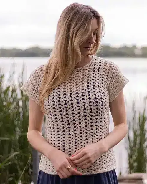 Woman wearing the Iris Stitch tee in a tan color with a navy blue dress underneath. The model is standing in front of a lake and looking off to the side. The photo shows off the mesh like stitch pattern of the tee.